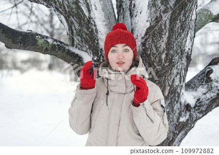 Portrait of attractive surprised girl. Pretty girl in red knitted hat and mittens under snow covered tree. Pleasant winter weather 109972882