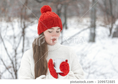 Young woman in red hat and mittens in snowy park. Beautiful girl with cup of beverage in winter outdoor 109972885
