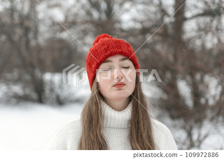 Portrait of young woman with closed eyes in woolen hat outdoors, cold weather. Young woman in knitted red hat and mittens, winter park Portrait of young woman with closed eyes in woolen hat outdoors, cold weather. Young woman in knitted red hat and mittens, winter park 109972886