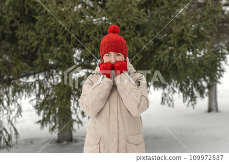 Young girl in red woolen hat and mittens warms hands standing near green pine tree in winter park or forest. Winter walk 109972887