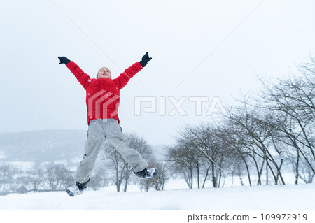 Happy teenage boy in red jacket jumps during snowfall. Child with his hands up and enjoys the winter holidays. Happy teenage boy in red jacket jumps during snowfall. Child with his hands up and enjoys the winter holidays. 109972919
