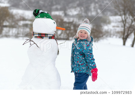 Smiling little child making a big snowman outdoor during winter holidays. Snowman is taller than child. Winter holidays. Smiling little child making a big snowman outdoor during winter holidays. Snowman is taller than child. Winter holidays. 109972983