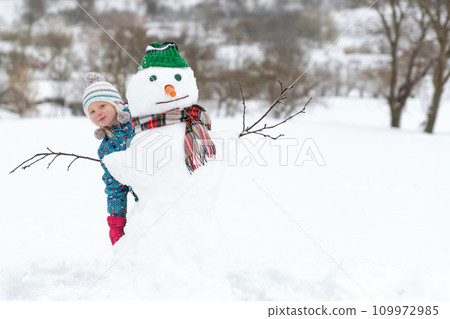 Happy child plays with snowman on winter walk. Kid outside building snow figure. Winter activities for kids. Happy child plays with snowman on winter walk. Kid outside building snow figure. Winter activities for kids. 109972985