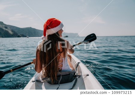Woman in kayak back view. Happy young woman in Santa hat floating in kayak on calm sea. Summer holiday vacation and cheerful female people relaxing having fun on the boat. Woman in kayak back view. Happy young woman in Santa hat floating in kayak on calm sea. Summer holiday vacation and cheerful female people relaxing having fun on the boat. 109973215