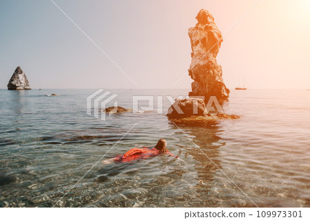Woman travel sea. Happy tourist in red dress enjoy taking picture outdoors for memories. Woman traveler posing in sea beach, surrounded by volcanic mountains, sharing travel adventure journey 109973301