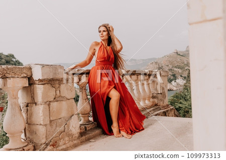 Woman travel portrait. Happy woman with long hair looking at camera and smiling. Close up portrait cute woman in a res long dress posing on backdrop of old travel city near sea 109973313