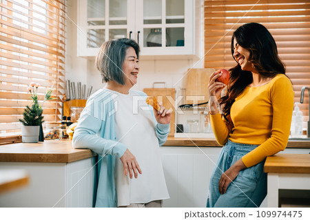 A smiling mother and her adult daughter, an attractive young woman, stand together in the kitchen, holding an apple. Their bond radiates happiness and family togetherness. A smiling mother and her adult daughter, an attractive young woman, stand together in the kitchen, holding an apple. Their bond radiates happiness and family togetherness. 109974475