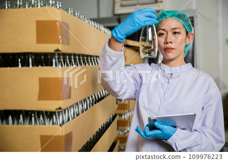 A QC woman working in a beverage factory utilizes a digital tablet to check products on the conveyor belt. She ensures quality control while examining the bottling line for liquid manufacturing. 109976223
