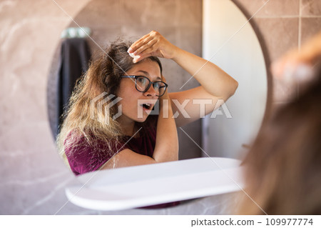 Gray haired surprised caucasian middle aged woman looking at grey hair head in mirror reflection. Early gray hair concept Gray haired surprised caucasian middle aged woman looking at grey hair head in mirror reflection. Early gray hair concept 109977774