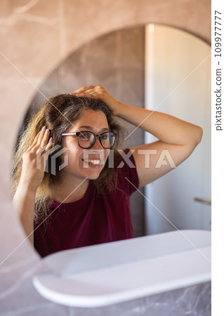 Gray haired surprised caucasian middle aged woman looking at grey hair head in mirror reflection. Early gray hair concept Gray haired surprised caucasian middle aged woman looking at grey hair head in mirror reflection. Early gray hair concept 109977777