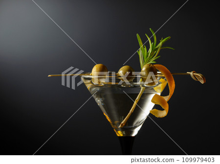 Classic dry martini cocktail with green olives, lemon peel, and rosemary on a black background. Classic dry martini cocktail with green olives, lemon peel, and rosemary on a black background. 109979403