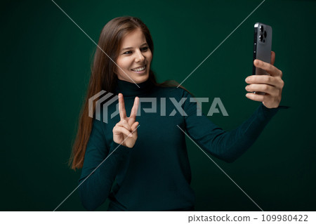 Caucasian young woman dressed in a green turtleneck takes a selfie on a smartphone Caucasian young woman dressed in a green turtleneck takes a selfie on a smartphone 109980422