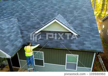 Using ladder, worker cleans debris, dirt, fallen leaves from clogged roof gutter drain Using ladder, worker cleans debris, dirt, fallen leaves from clogged roof gutter drain 109980960