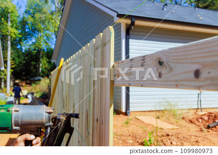 With use of an air nail gun man constructs sections of wooden fence in his yard from planks 109980973