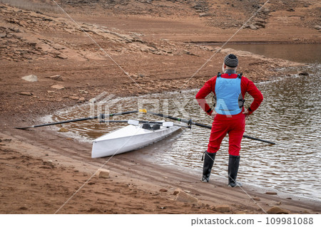 Senior male rower wearing drysuit with a coastal rowing shell on a shore of Horsetooth Reservoir in fall or winter scenery. 109981088