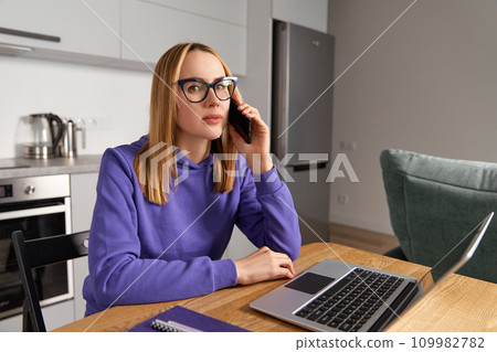 Young adult woman in violet hoodie speaks on a cell phone and using laptop. Work from home concept 109982782