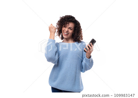 young caucasian woman with curly dark hair dressed in a casual warm blue jacket holds a smartphone 109983147