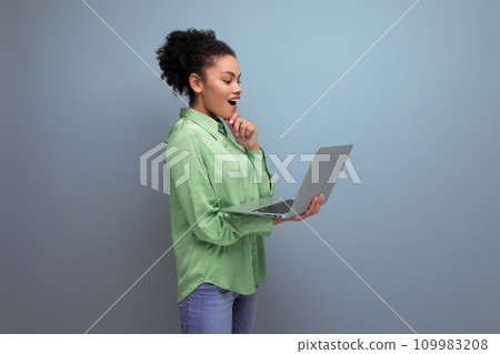 young brunette hispanic woman with curly hair in green rejoices holding a laptop in her hand against young brunette hispanic woman with curly hair in green rejoices holding a laptop in her hand against 109983208