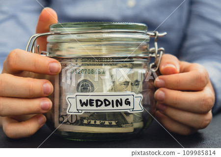 Female hands holding Glass jar full of American currency dollars cash banknote with text WEEDING. Preparation saving money. Moderate consumption and economy Female hands holding Glass jar full of American currency dollars cash banknote with text WEEDING. Preparation saving money. Moderate consumption and economy 109985814