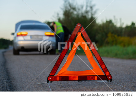 Unrecognizable sad driver in reflective vest. Male driver standing near broken car with open up hood. Red triangle to warn other road users of car breakdown or engine failure stop at countryside Unrecognizable sad driver in reflective vest. Male driver standing near broken car with open up hood. Red triangle to warn other road users of car breakdown or engine failure stop at countryside 109985832