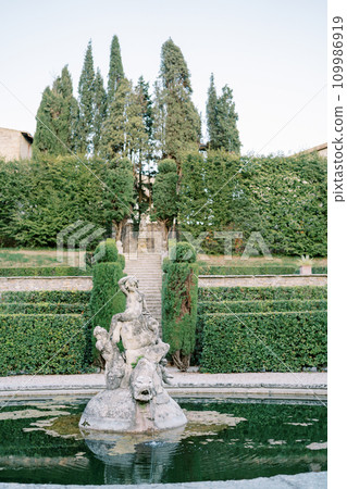 Ancient stone fountain in a green garden with a nymph sitting on a large fish 109986919