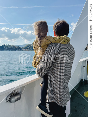 Dad with a little girl in his arms stands on board a boat floating on the sea and looks at the shore 109986985