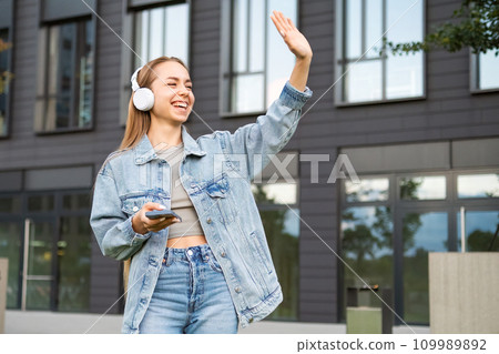 Young woman with a smile and headphones waves to someone on the street.  109989892