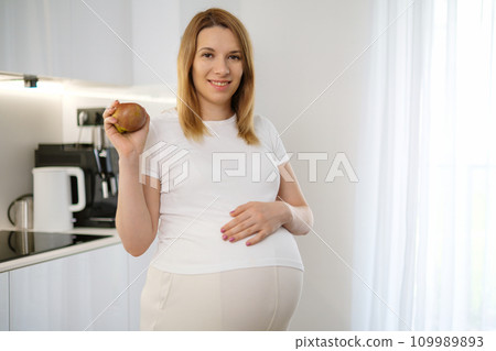 A smiling blonde hair pregnant woman is holding an apple and putting her hand on her belly in the kitchen. Diet during pregnancy.  109989893
