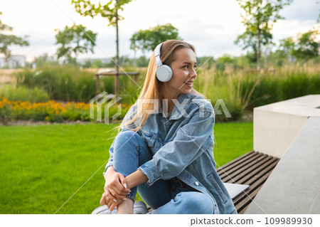 Young lady is seated with legs folded on a bench, engrossed in music through her headphones in the park  109989930