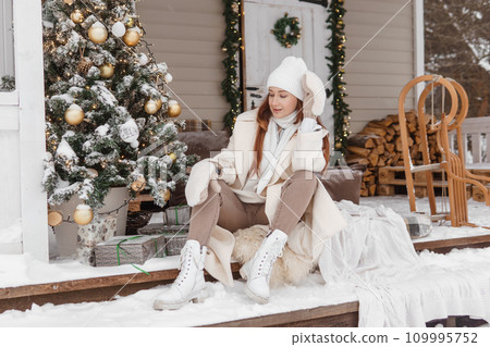 A woman in white winter clothes on the veranda of a country house decorated for the Christmas holidays. Wooden house. A woman in white winter clothes on the veranda of a country house decorated for the Christmas holidays. Wooden house. 109995752
