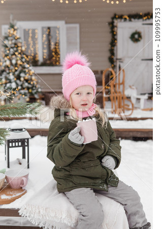 A little girl in winter clothes with a cup of hot cocoa with marshmallows, in the courtyard of a country house decorated for the Christmas holidays. Christmas holidays. 109995753
