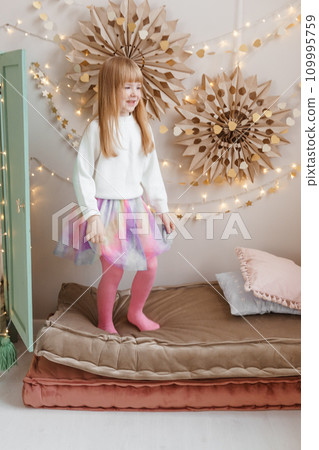 A little red-haired girl jumps on mattresses in a nursery decorated for the Christmas holidays. 109995759