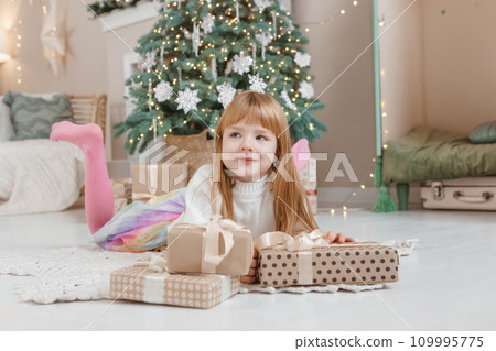 A little red-haired girl lies next to Christmas gifts in a craft package. Happy Christmas concept. A little red-haired girl lies next to Christmas gifts in a craft package. Happy Christmas concept. 109995775