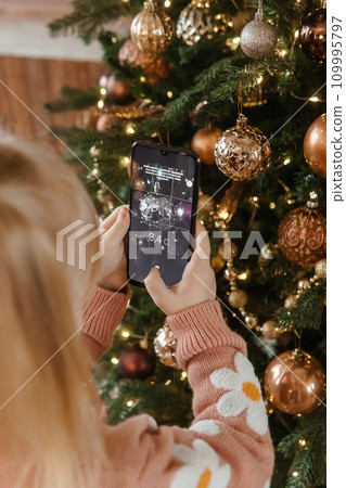 A little blonde girl photographs balloons on a Christmas tree in a festive interior decorated in a New Year's style. The concept of a merry Christmas. A little blonde girl photographs balloons on a Christmas tree in a festive interior decorated in a New Year's style. The concept of a merry Christmas. 109995797