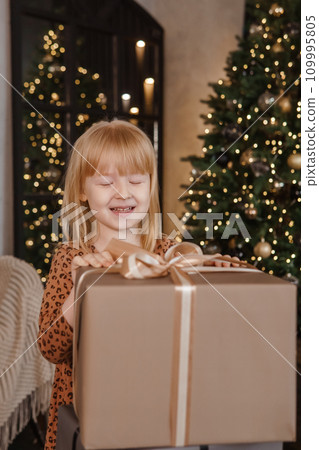 A little blonde girl in a festive interior decorated in a New Year's style. A child holds a Christmas gift in a craft package. A little blonde girl in a festive interior decorated in a New Year's style. A child holds a Christmas gift in a craft package. 109995805