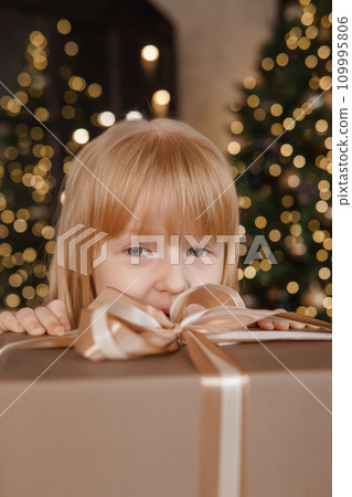 A little blonde girl in a festive interior decorated in a New Year's style. A child holds a Christmas gift in a craft package. A little blonde girl in a festive interior decorated in a New Year's style. A child holds a Christmas gift in a craft package. 109995806