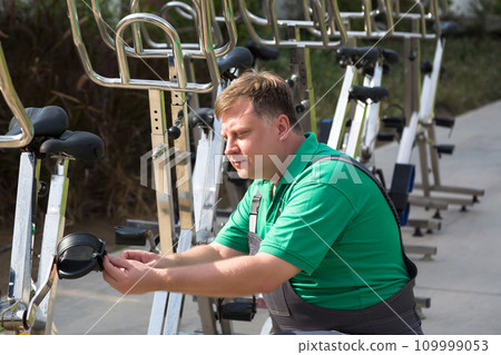 A man in overalls repairs and maintains sports bicycle simulators A man in overalls repairs and maintains sports bicycle simulators 109999053