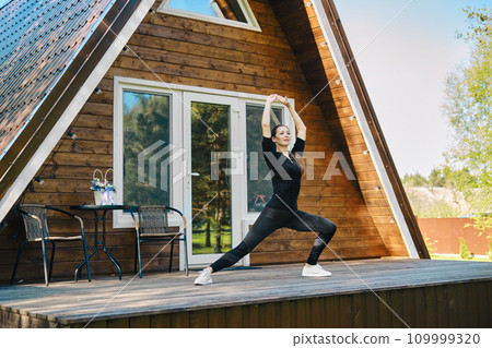 Sporty woman in sport clothing does stretching exercise on the terrace of wooden cabin 109999320