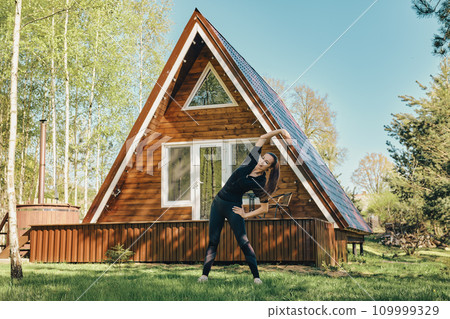 Sporty woman does side body bent on lawn of a log cabin Sporty woman does side body bent on lawn of a log cabin 109999329