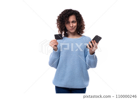 pretty young curly brunette woman dressed in a blue sweater holds a plastic bank card and smartphone 110000246