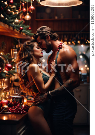 Intimate couple in a festive kitchen, with a Christmas tree and lights. Man shirtless, woman in bra, close by counter with fruit bowl. Cozy, affectionate atmosphere. 110003293