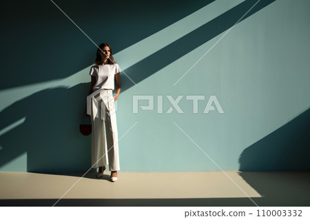 Elegant model in a sleek, monochromatic outfit, standing against a pastel wall, her shadow creating a sharp contrast on the minimalist background 110003332