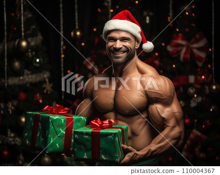 Muscular bodybuilder smiles and holding a gift box with bow. Wearing shirtless and Santa hat, beside Christmas tree with colorful baubles. Warm, cheerful holiday atmosphere. 110004367