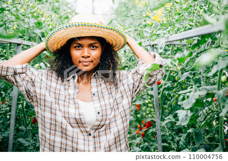 Woman farmer smiling standing in a greenhouse among tomato plants holding a rake. Portrait of cheerful adult owner caring for vegetable growth. Happiness in horticulture outdoors. Sunlight beauty is. Woman farmer smiling standing in a greenhouse among tomato plants holding a rake. Portrait of cheerful adult owner caring for vegetable growth. Happiness in horticulture outdoors. Sunlight beauty is. 110004756