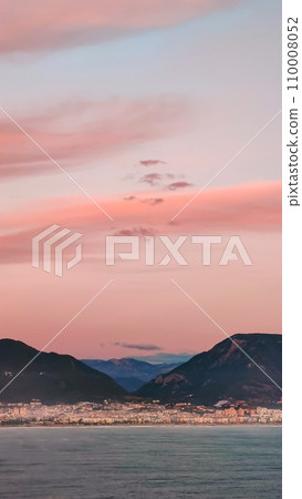 Pink clouds in evening sky over blue sea on background of city and mountains on horizon, vertical frame. 110008052