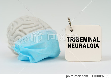 On a white surface there is a brain with a blue mask and a notepad with the inscription - Trigeminal neuralgia On a white surface there is a brain with a blue mask and a notepad with the inscription - Trigeminal neuralgia 110009233