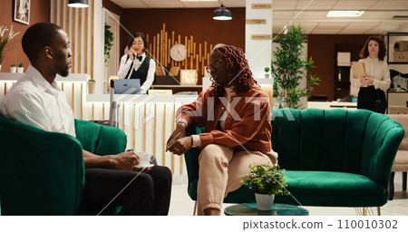 Hotel manager checking in with waiting guests, asking about any required hotel extra amenities or room upgrades. Tourists discussing booking options while bellboy picks up their baggage 110010302