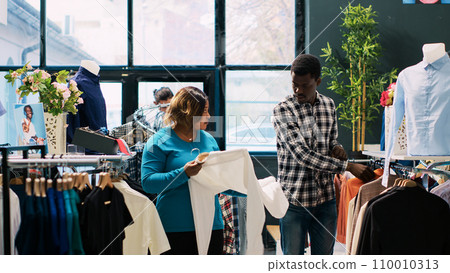 African american couple looking at fashionable clothes, checking items material in clothing store. Cheerful clients enjoying shopping session, buying stylish merchandise and accessories in modern 110010313