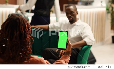 Company partners away on business trip waiting in hotel foyer to be checked in, holding chroma key green screen mock up smartphone. Coworkers chatting in cozy resort lobby, checking info on phone 110010314
