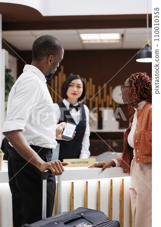 Young man paying for booking with mobile phone nfc, talking to receptionist about luxury resort services. Tourists making payment at pos terminal, filling in registration forms. 110010358